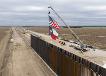 A crane lifts the US and Texas flags beside a border wall under construction, with vehicles and workers nearby and open fields on both sides.
