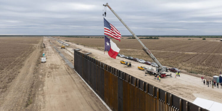 A crane lifts the US and Texas flags beside a border wall under construction, with vehicles and workers nearby and open fields on both sides.