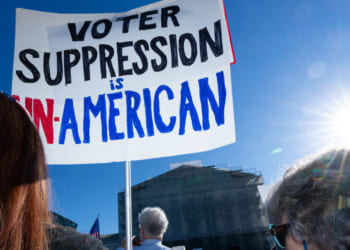 People stand outside in sunlight holding a sign that reads, Voter suppression is un-American, with part of the sign in red, black, and blue letters. A government building is visible in the background.