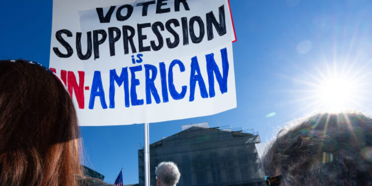 People stand outside in sunlight holding a sign that reads, Voter suppression is un-American, with part of the sign in red, black, and blue letters. A government building is visible in the background.