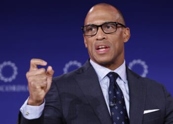 A man in a suit and glasses speaks while gesturing with his hand. He is positioned in front of a blue background with circular logos.