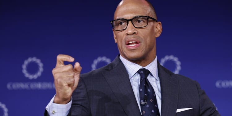 A man in a suit and glasses speaks while gesturing with his hand. He is positioned in front of a blue background with circular logos.