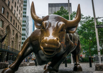 A close-up view of the Charging Bull bronze sculpture in New York Citys Financial District, with buildings and street signs visible in the background.