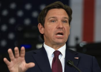 A man in a dark suit and red tie gestures while speaking, with an American flag in the blurred background.