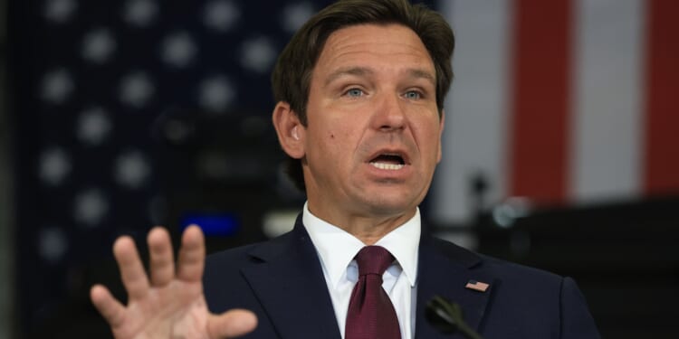 A man in a dark suit and red tie gestures while speaking, with an American flag in the blurred background.