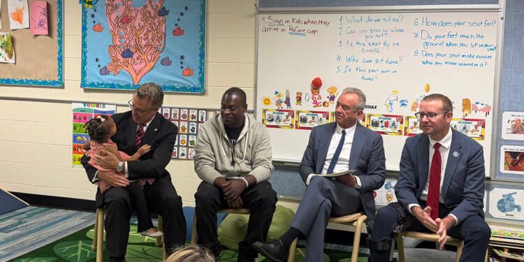 Four HHS officials sit in a classroom, one holding a dog. Behind them are colorful bulletin boards, childrens art, and classroom instructions displayed on the wall.
