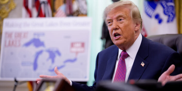 A man in a suit gestures while speaking in an office. Behind him is a blurred map labeled The Great Lakes and some United States flags.