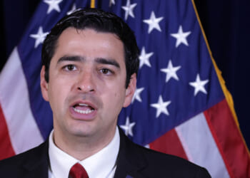 Gabe Evans in a suit and red tie speaks in front of two American flags, appearing to address an audience or the media.