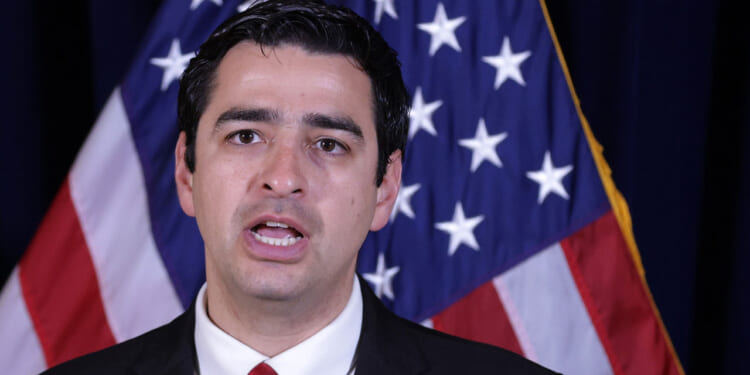 Gabe Evans in a suit and red tie speaks in front of two American flags, appearing to address an audience or the media.