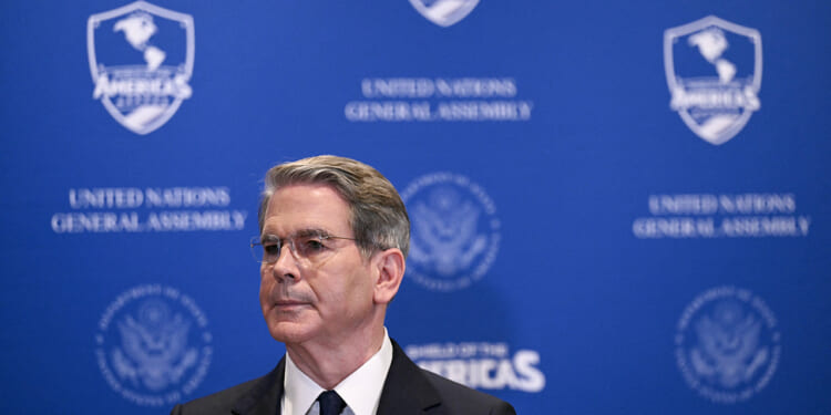 A man in a suit and glasses stands in front of a blue backdrop with United Nations General Assembly and U.S. logos, looking to the side.