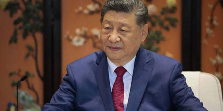 A man in a blue suit and red tie sits before a decorative floral painted screen in orange and green tones, with a microphone visible to his left.