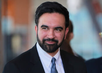 A man with dark hair and a beard, wearing a black suit, white shirt, and blue tie, is smiling slightly while looking to the side. Another person in the background is blurred and wearing sunglasses.