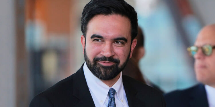 A man with dark hair and a beard, wearing a black suit, white shirt, and blue tie, is smiling slightly while looking to the side. Another person in the background is blurred and wearing sunglasses.