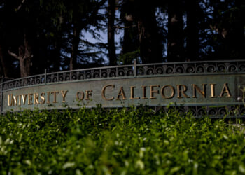 A metal sign reads University of California in gold letters, partially obscured by green foliage, with trees and a dark background behind it.