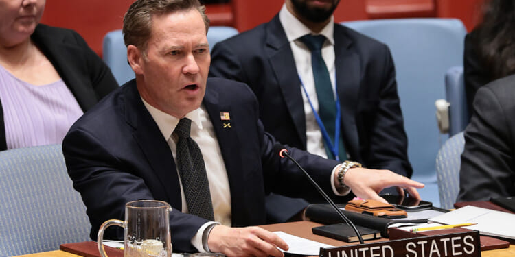 Mike Walz in a suit speaks emphatically at a conference table with a sign reading United States. He has papers and a microphone in front of him, and people are seated nearby.