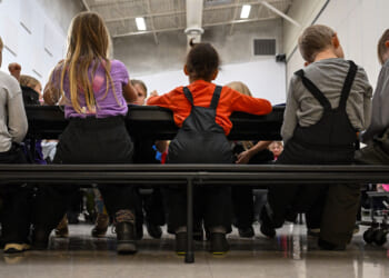 A group of children wearing winter clothes sit closely together on benches at a table in a brightly lit room, seen from behind.