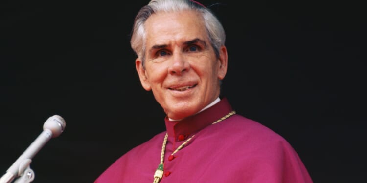 An older man in religious attire with a red robe and gold chain stands at a microphone, smiling slightly, against a dark background.