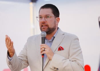 A man in a light blazer and glasses speaks into a microphone, gesturing with his left hand. He wears a red pocket square and stands in front of a blurred background.