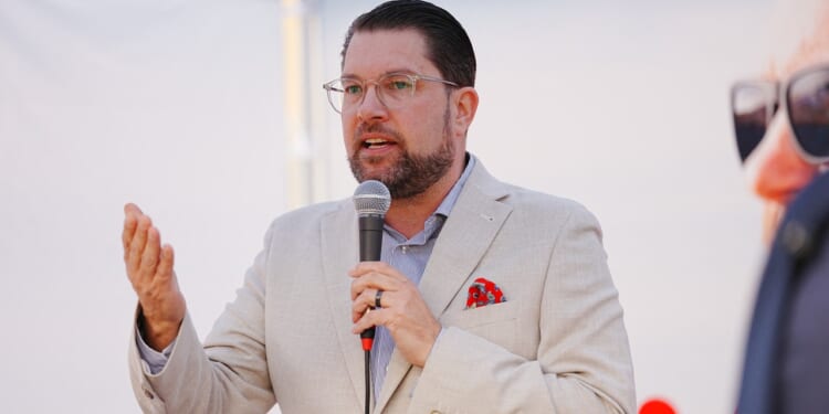 A man in a light blazer and glasses speaks into a microphone, gesturing with his left hand. He wears a red pocket square and stands in front of a blurred background.