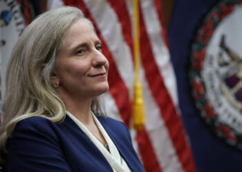 A woman with long gray hair in a blue blazer stands in front of U.S. and Virginia state flags, looking to the side and smiling slightly.