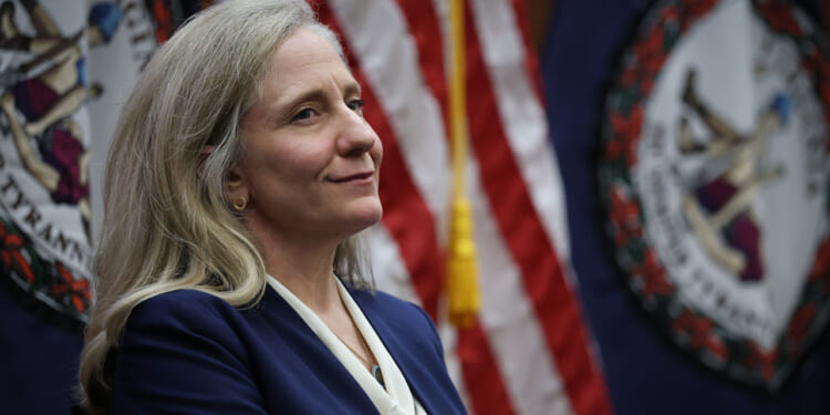 A woman with long gray hair in a blue blazer stands in front of U.S. and Virginia state flags, looking to the side and smiling slightly.