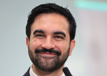 A man with dark hair and a beard smiles at the camera, wearing a suit and standing in front of a blurred light background.