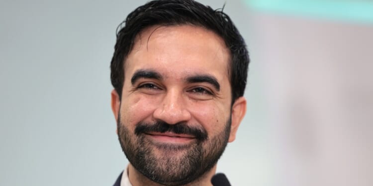 A man with dark hair and a beard smiles at the camera, wearing a suit and standing in front of a blurred light background.