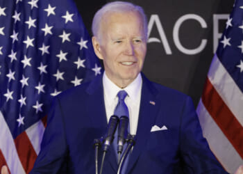 Joe Biden in a blue suit speaks at a podium with microphones, standing between two American flags, with a black background featuring partial white text.