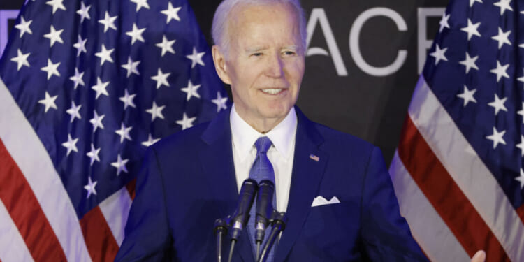 Joe Biden in a blue suit speaks at a podium with microphones, standing between two American flags, with a black background featuring partial white text.