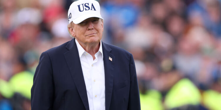 Donald Trump wearing a white USA cap, dark jacket, and white shirt stands outdoors with a blurred crowd in the background.