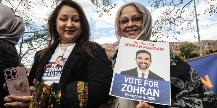 Two women outdoors hold a Vote for Zohran campaign sign and a phone. One wears a shirt with the candidates photo; both are smiling. There are trees and a blue sky in the background.
